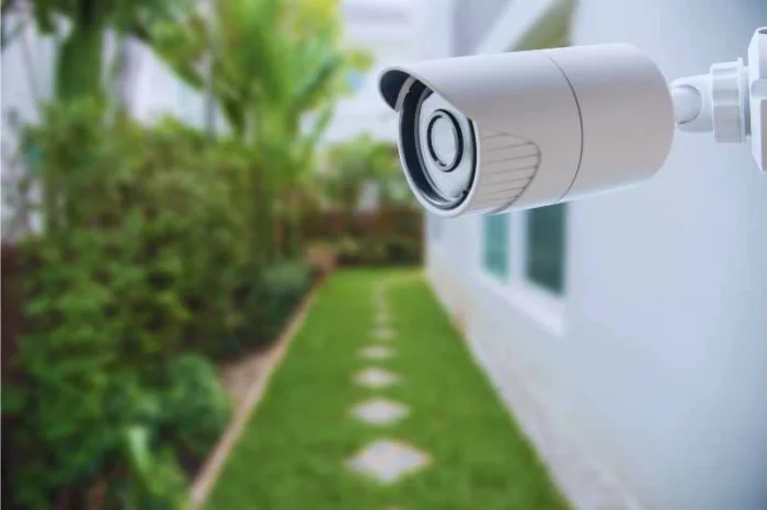 White commercial outdoor security camera mounted on the side of a one-story house by a green lawn.