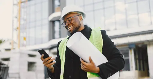 developer wearing reflective vest and hard hat while holding blueprint and smiling at phone in front of building