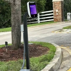 Side view of ButterflyMX intercom displaying welcome screen, mounted on a pedestal at vehicle entrance of gated community Side view of ButterflyMX intercom displaying welcome screen, mounted on a pedestal at vehicle entrance of gated community